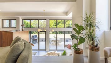 Una sala de estar moderna con un sofá, dos plantas en macetas, una zona de comedor con una mesa y sillas, y vistas a un patio exterior a través de puertas de cristal amplias.
