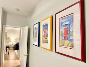 Ein Flur mit gerahmten Kunstwerken an der Wand, eine zurueckliegende Tuere zu einem Zimmer und ein sichtbares Badezimmer mit Toilette und Waschbecken.