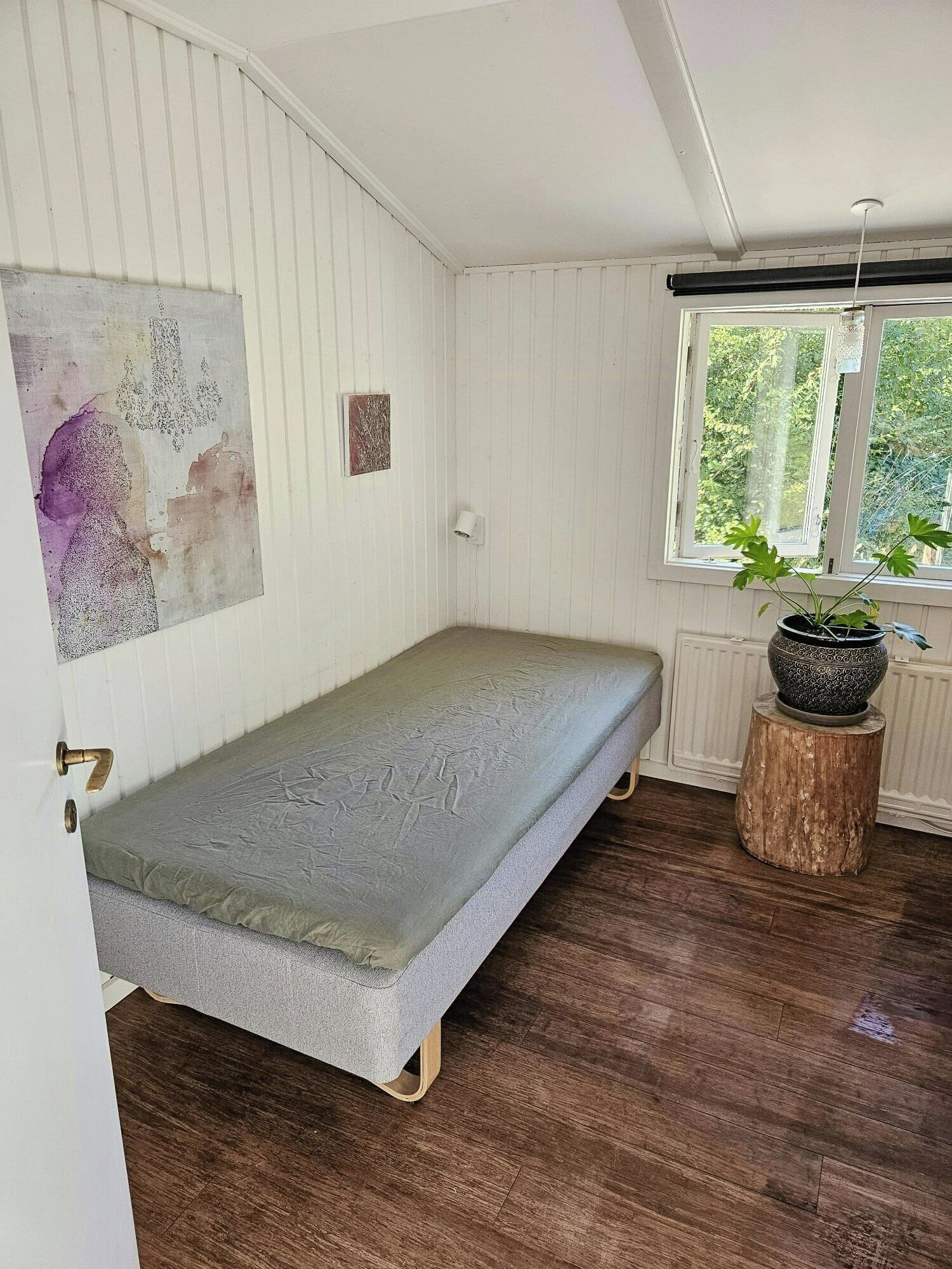 A single bed in a room with white walls, a window with a view of greenery, and a small potted plant on a wooden stump.