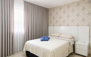 A bedroom with a bed, curtains, a nightstand, and a wall with a floral pattern. - Apartamento Fiji c/ Piscina - Bal. Picarras/sc (Picarras)