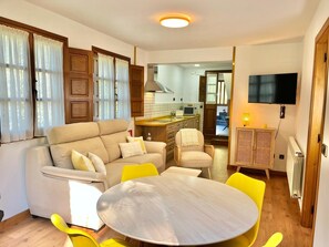 Una sala de estar con un sofá, sillas amarillas, una mesa redonda, un televisor y una cocina al fondo.