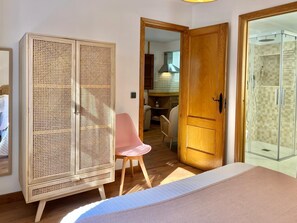A bedroom with a bed, a wooden wardrobe, a pink chair, a wooden door, and a glass shower enclosure.