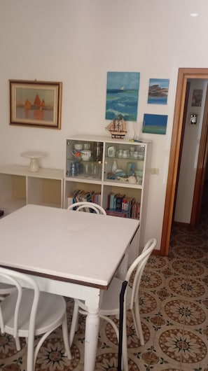 A dining area with a white table and chairs, a glass display cabinet with books, and a wall with framed pictures. - Casa Glicine Isola Del Giglio: Sea View, Private Terrace and Garden (Giglio Campese)