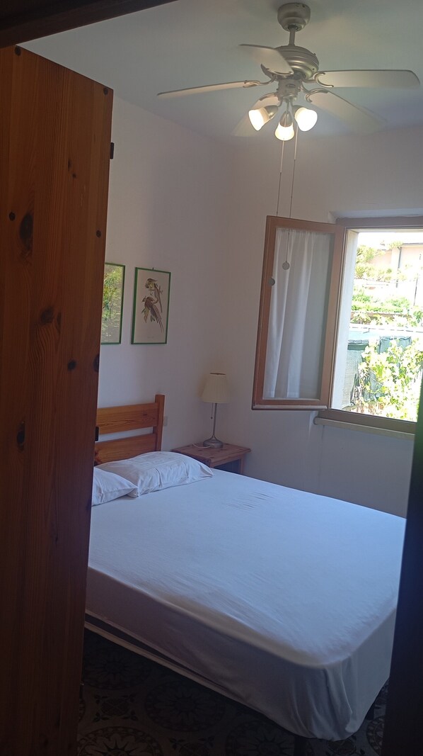 A bedroom with a bed, a ceiling fan, a window with curtains, and framed pictures on the wall. - Casa Glicine Isola Del Giglio: Sea View, Private Terrace and Garden (Giglio Campese)