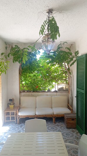 A cozy room with a sofa, a small table, a wicker chair, and a window with a view of greenery. - Casa Glicine Isola Del Giglio: Sea View, Private Terrace and Garden (Giglio Campese)