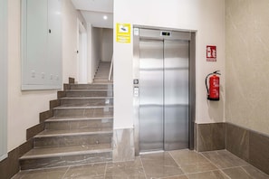 Eine Hotelhalle mit Treppe, Aufzug, Feuerlöscher und Schild, das eine rund um die Uhr verfügbare Aufzugsanbindung anzeigt.
