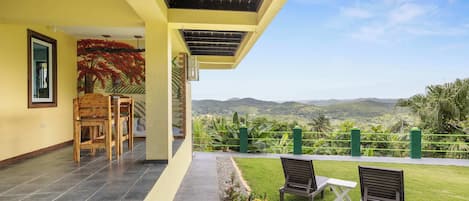 A covered outdoor area with a view of hills, a wooden bench, a table, and a chair.
