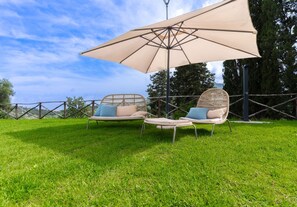 Ein Sitzbereich im Freien mit zwei Korbstühlen und einem großen Sonnenschirm, gelegen auf einer grünen Rasenfläche mit Bäumen im Hintergrund.