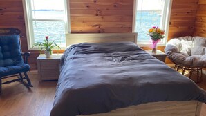 Una habitación de cabaña de madera con cama, una silla, una mesita de noche y ventana con vistas al agua.
