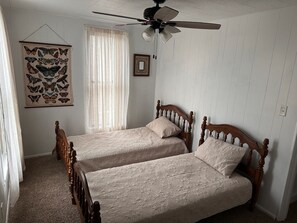 Ein Schlafzimmer mit zwei hölzernen Betten, einem Deckenventilator, einem Fenster mit durchsichtigen Vorhängen und Wandbild mit einem Schmetterling.