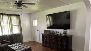 A living room with a wooden floor, a dark wooden cabinet, a vintage trunk, a ceiling fan, and a flat-screen TV mounted on the wall.
