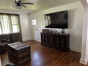 A living room with a wooden floor, a dark wooden cabinet, a vintage trunk, a ceiling fan, and a flat-screen TV mounted on the wall.