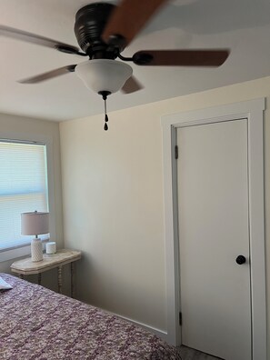 Ein Schlafzimmer mit Deckenventilator, einem Bett mit gemustertem Bettbezug, einem Nachttisch mit Lampe und einem Fenster mit Jalousien.