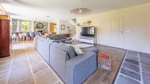 A spacious living room with a large grey sofa, a television, and a stone fireplace. - Le Domaine du Rocher - Pour 12 avec grand jardin (Neussargues-Moissac)