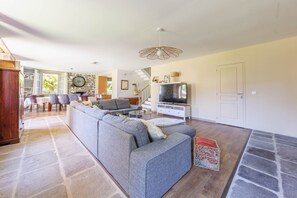 A spacious living room with a large grey sofa, a television, and a stone fireplace. - Le Domaine du Rocher - Pour 12 avec grand jardin (Neussargues-Moissac)