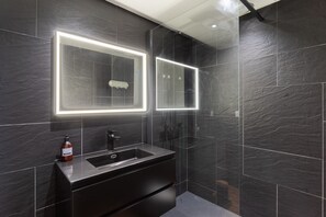 Ein modernes Badezimmer mit dunklen Fliesen, einem Waschbecken und zwei beleuchteten Spiegeln.
