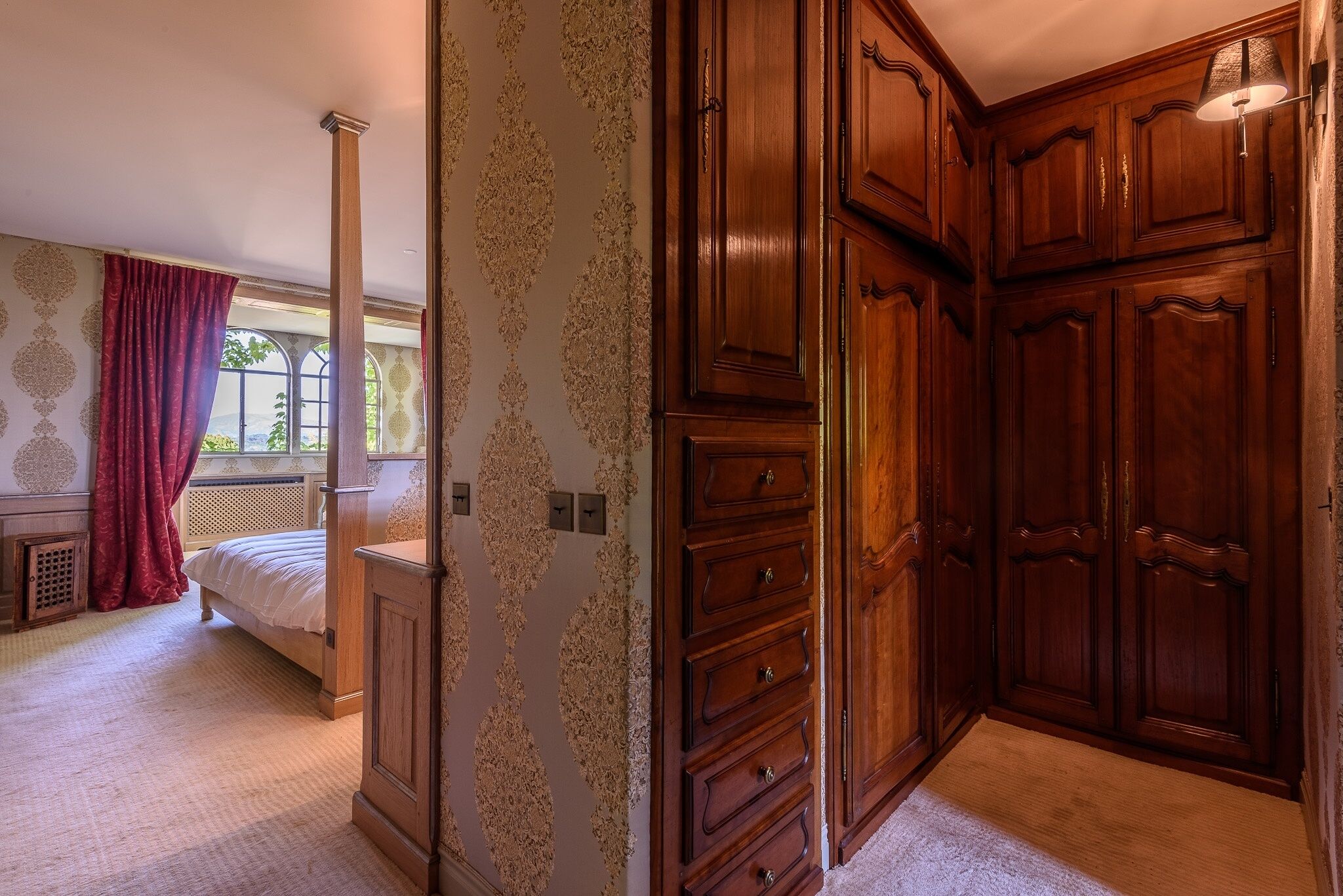 A bedroom with a large wooden wardrobe, a bed with a red curtain, and a window with a view.