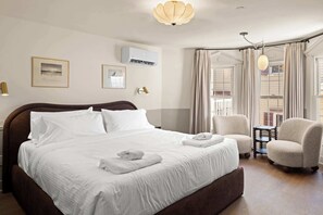 Una cama bien hecha con sábanas y toallas blancas, un aire acondicionado montado en la pared, cuadros enmarcados y un candelabro.
