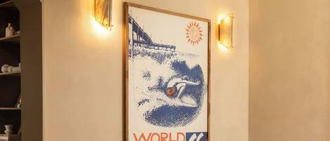 A room with a framed poster of the World Surfing Championships, two upholstered chairs, a wooden floor, and a chandelier.