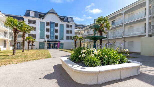 A courtyard with a fountain, palm trees, and a building with balconies. - Green Flat - Appt proche des Sables d'Olonnes (L'Aiguillon-sur-Vie)