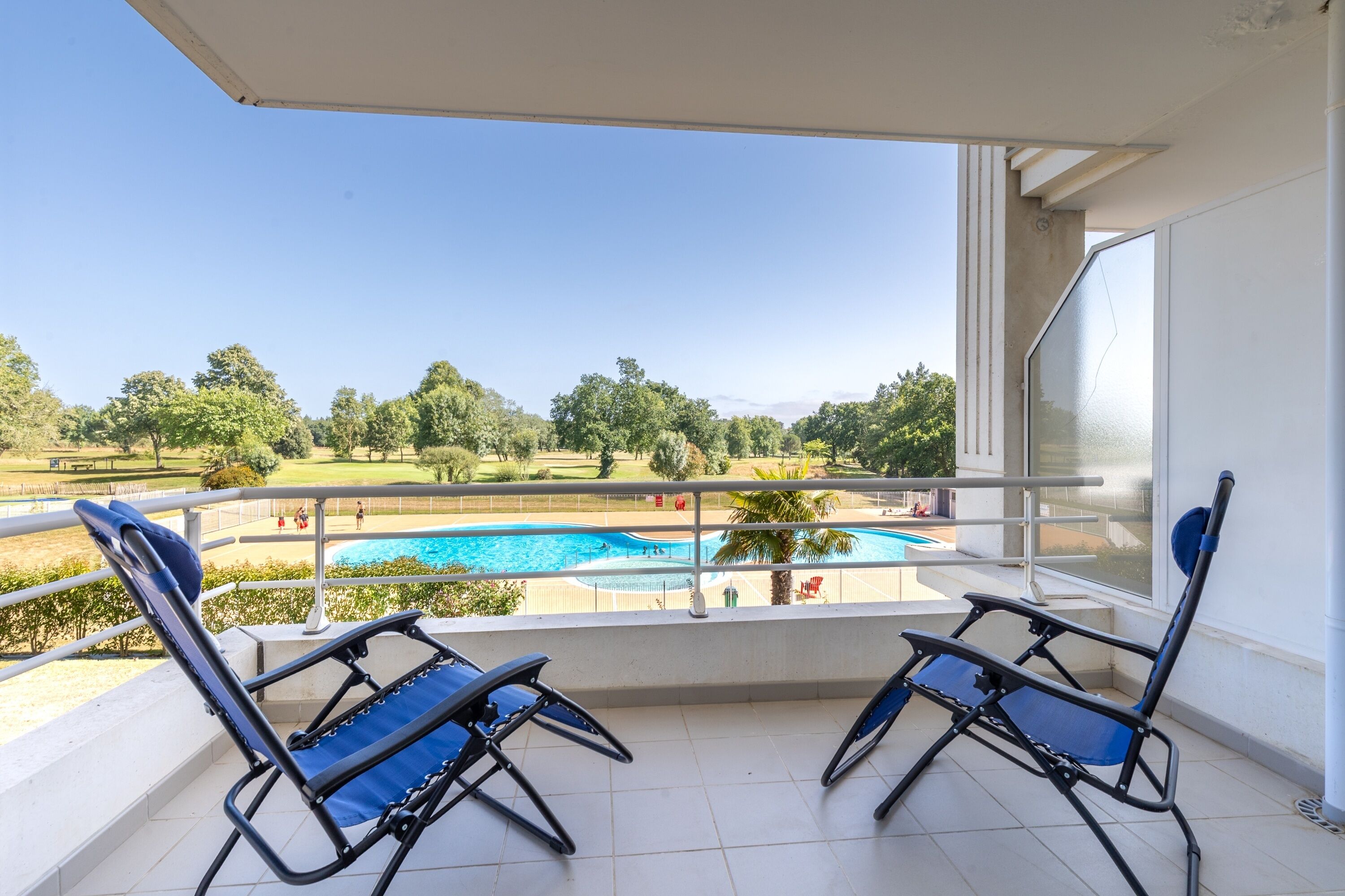 A balcony with two lounge chairs, a pool, and a view of a golf course.