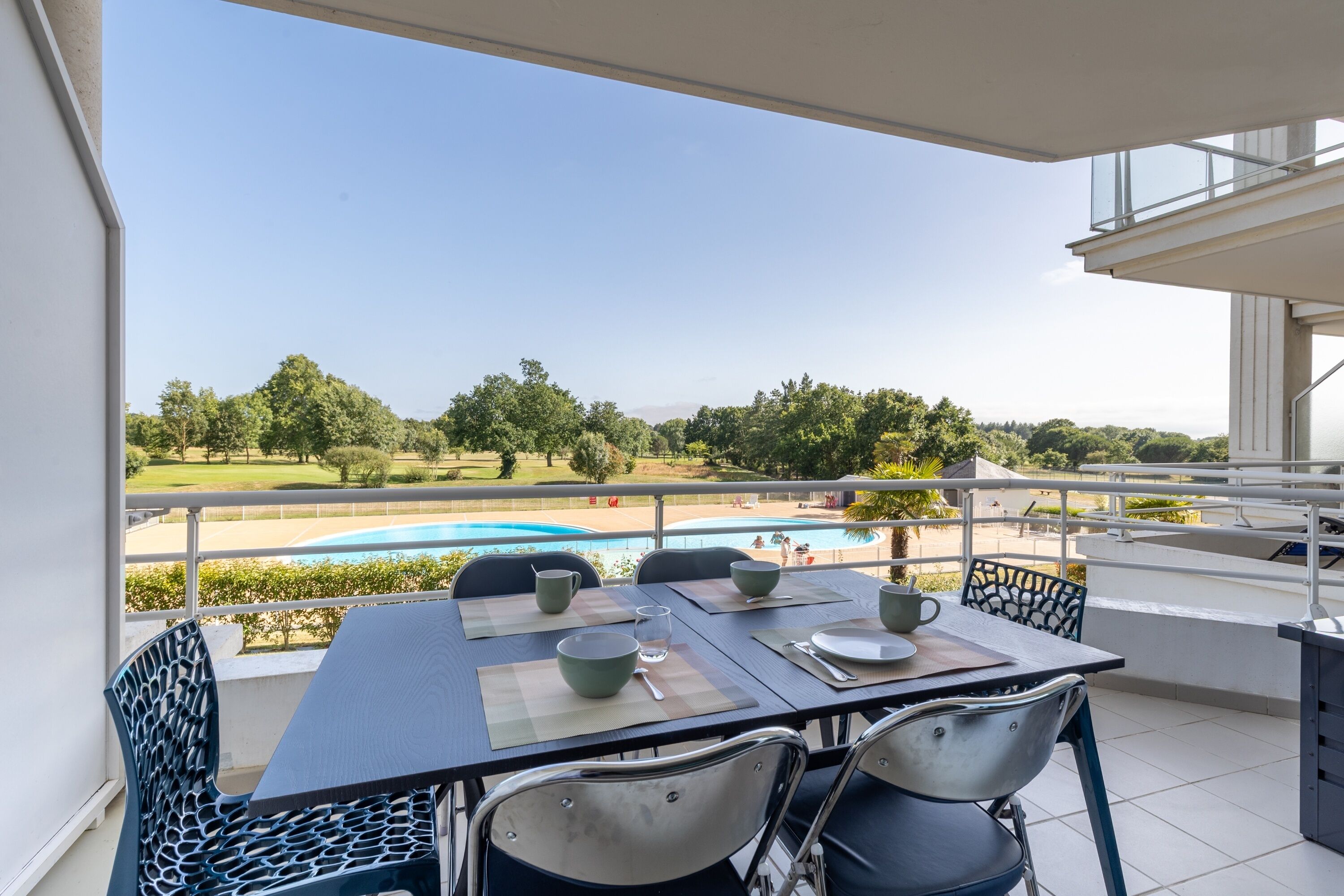 Un balcón con mesa dispuesta para cuatro, con vistas a una piscina y follaje.