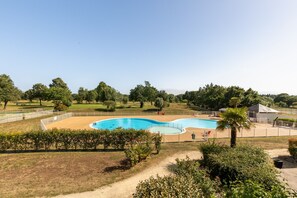 Ein Schwimmbecken mit Holzterrasse, umgeben von Grünflächen und blauem Himmel.