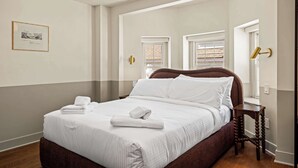 Un letto rifatto con lenzuola e asciugamani bianchi, una testiera in legno e un comodino in legno con una lampada.