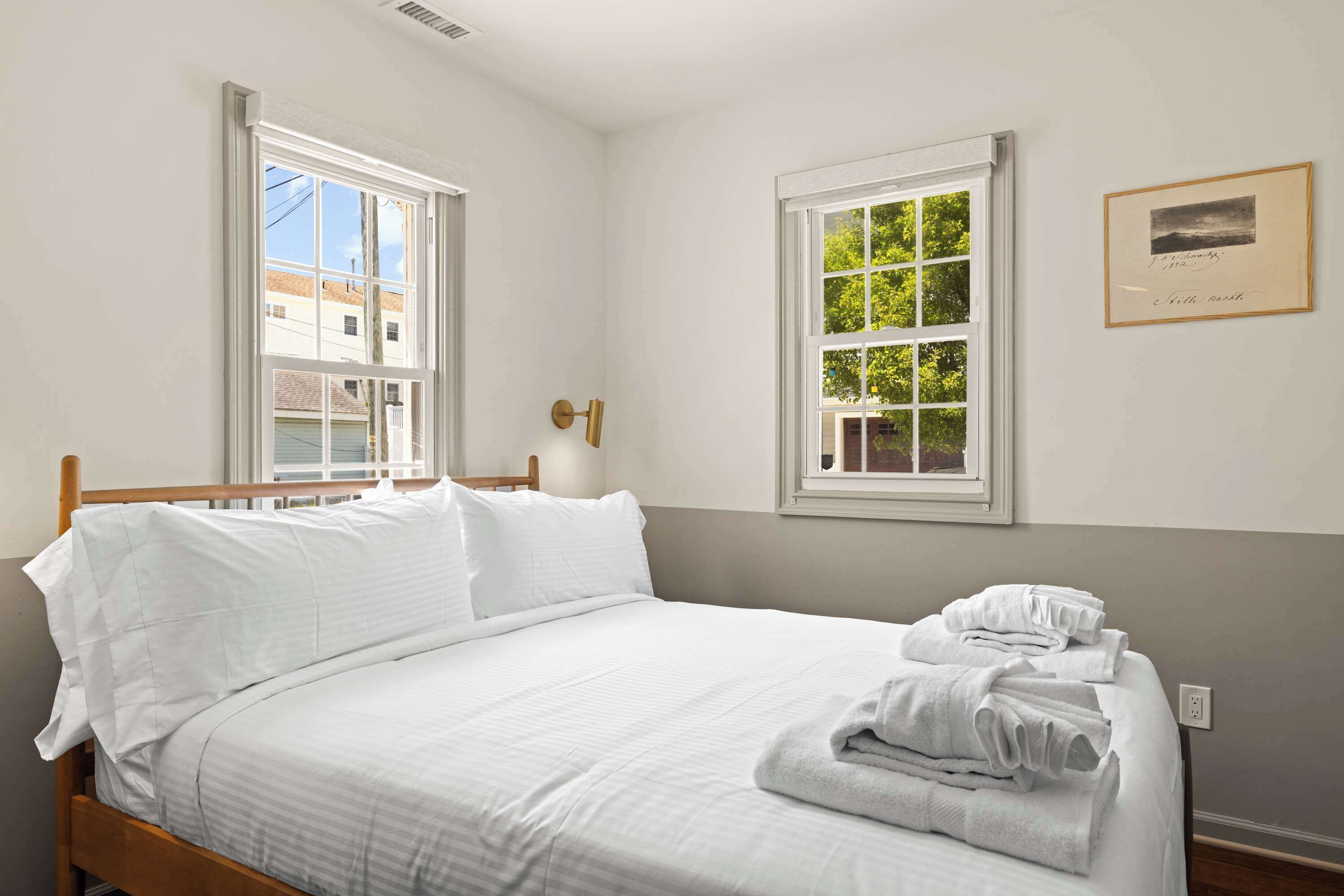 A neatly made bed with white linens and towels, two windows with white frames, and a framed picture on the wall.