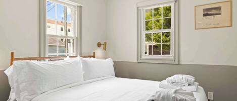 A neatly made bed with white linens and towels, two windows with white frames, and a framed picture on the wall.