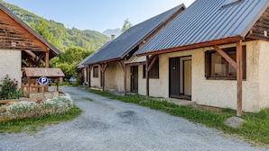 A rustic building with a gravel driveway, wooden porch, and a mountain backdrop. - Le Pré de l'Arche - À 400 m des télécabines (Oz)
