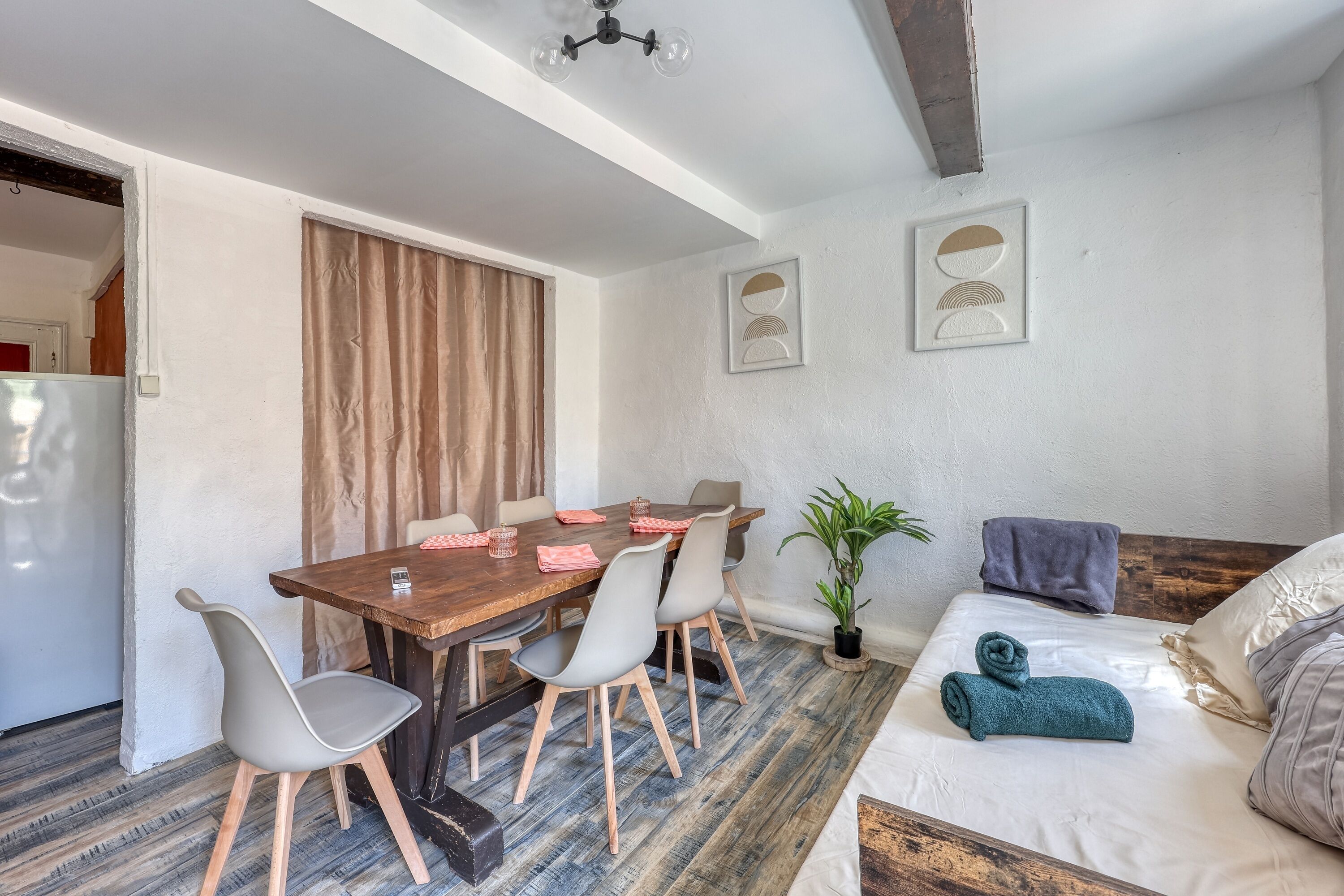 Ein Zimmer mit einem Holztisch, grauen Stühlen, einem Sofa, einer Topfpflanze und einem Bett mit einem zusammengerollten Handtuch.