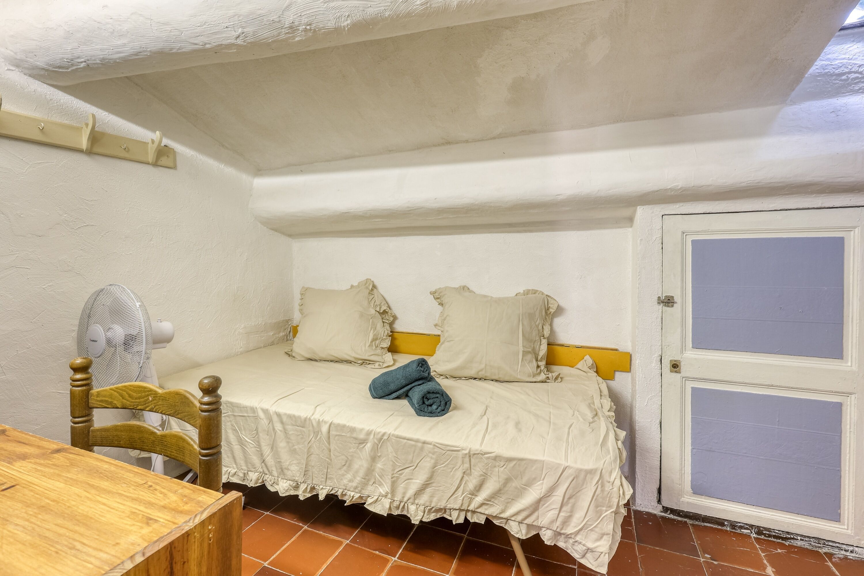Een kleine, knusse kamer met een bed, een ventilator, een houten stoel en een deur.