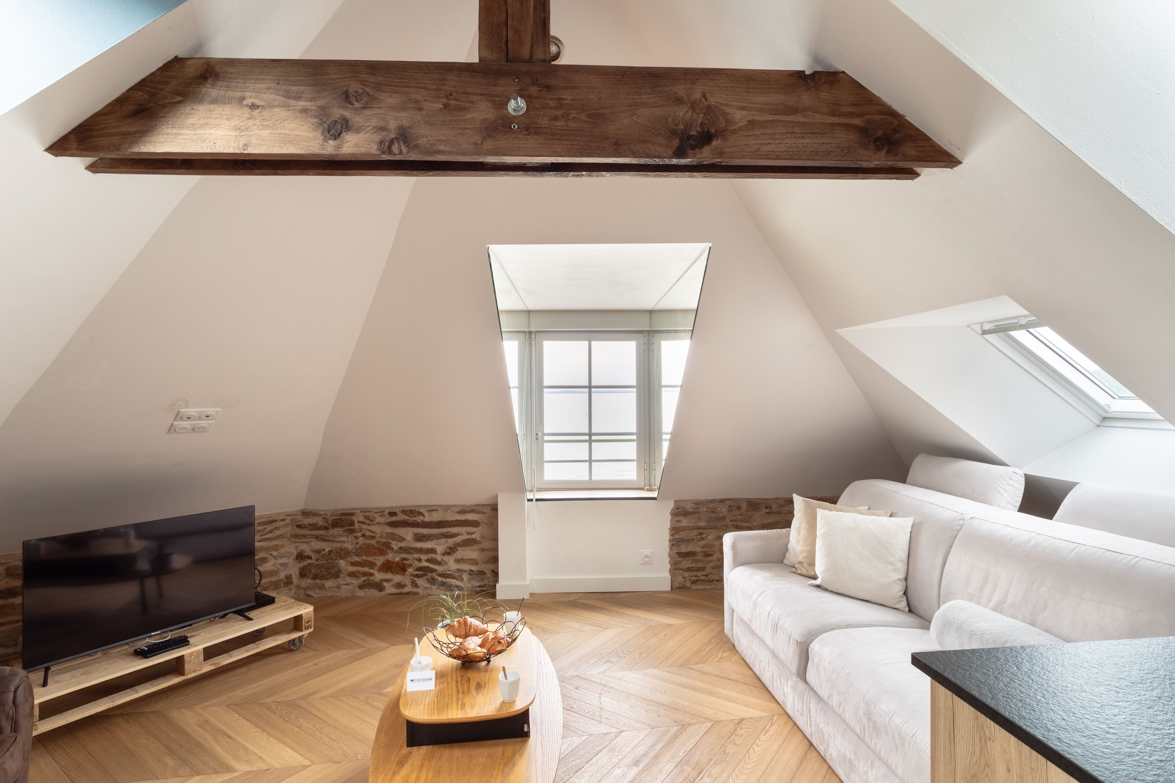 A loft-style room with a sloped ceiling, a large window, a flat-screen TV, a wooden coffee table, and a white sofa.