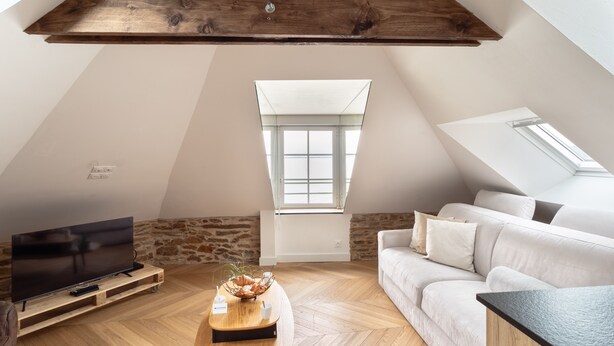 A loft-style room with a sloped ceiling, a large window, a flat-screen TV, a wooden coffee table, and a white sofa. - La Houle - Bel appartement avec vue mer (Cancale)
