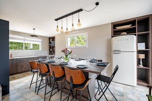 A modern kitchen with a central island, wooden chairs, a white refrigerator, and a window with a view of greenery. - Les Volets Bleus de Rhuys - Avec piscine privée (Saint-Gildas-de-Rhuys)