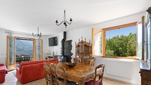 A dining area with a wooden table and chairs, a red sofa, and a view of greenery through large windows. - Le Nid de Ciambonson - Terrasse avec vue montagne (La Tour)