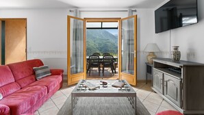 A living room with a red sofa, a coffee table, a TV, and a view of a mountain through a glass door. - Le Nid de Ciambonson - Terrasse avec vue montagne (La Tour)