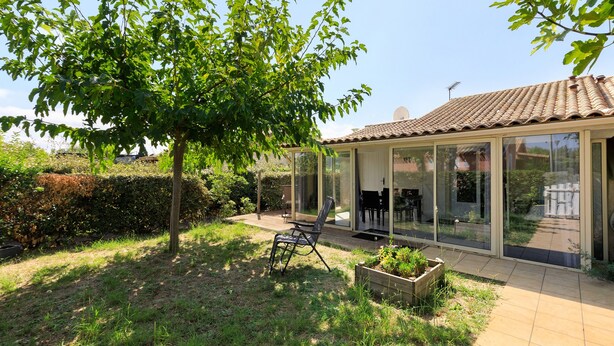 A covered outdoor seating area with a table and chairs, surrounded by greenery and a building with a tiled roof.