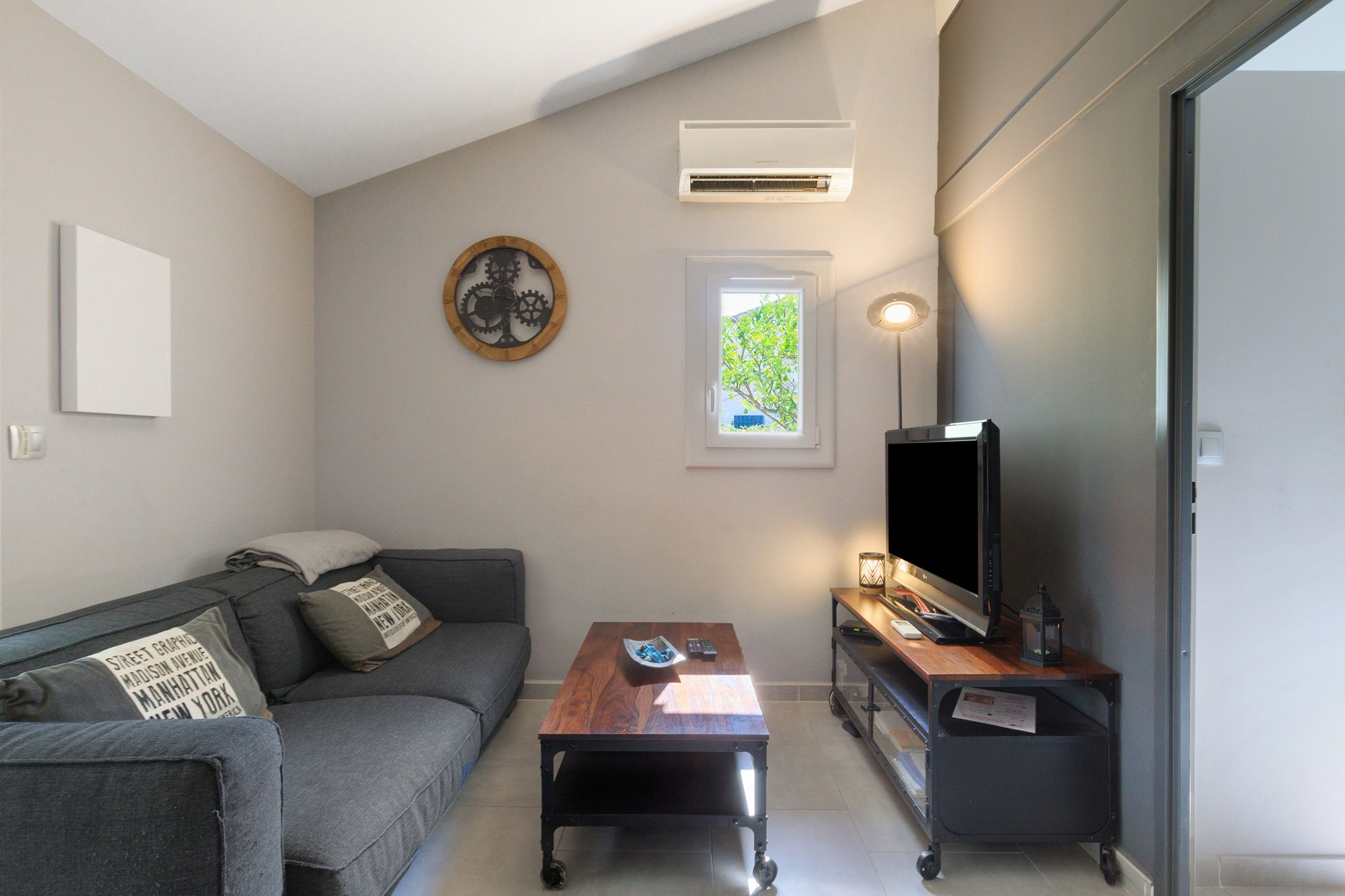 Een moderne woonkamer met een grijze bank, een houten salontafel, een flatscreen televisie op een standaard en een raam met uitzicht op groen.