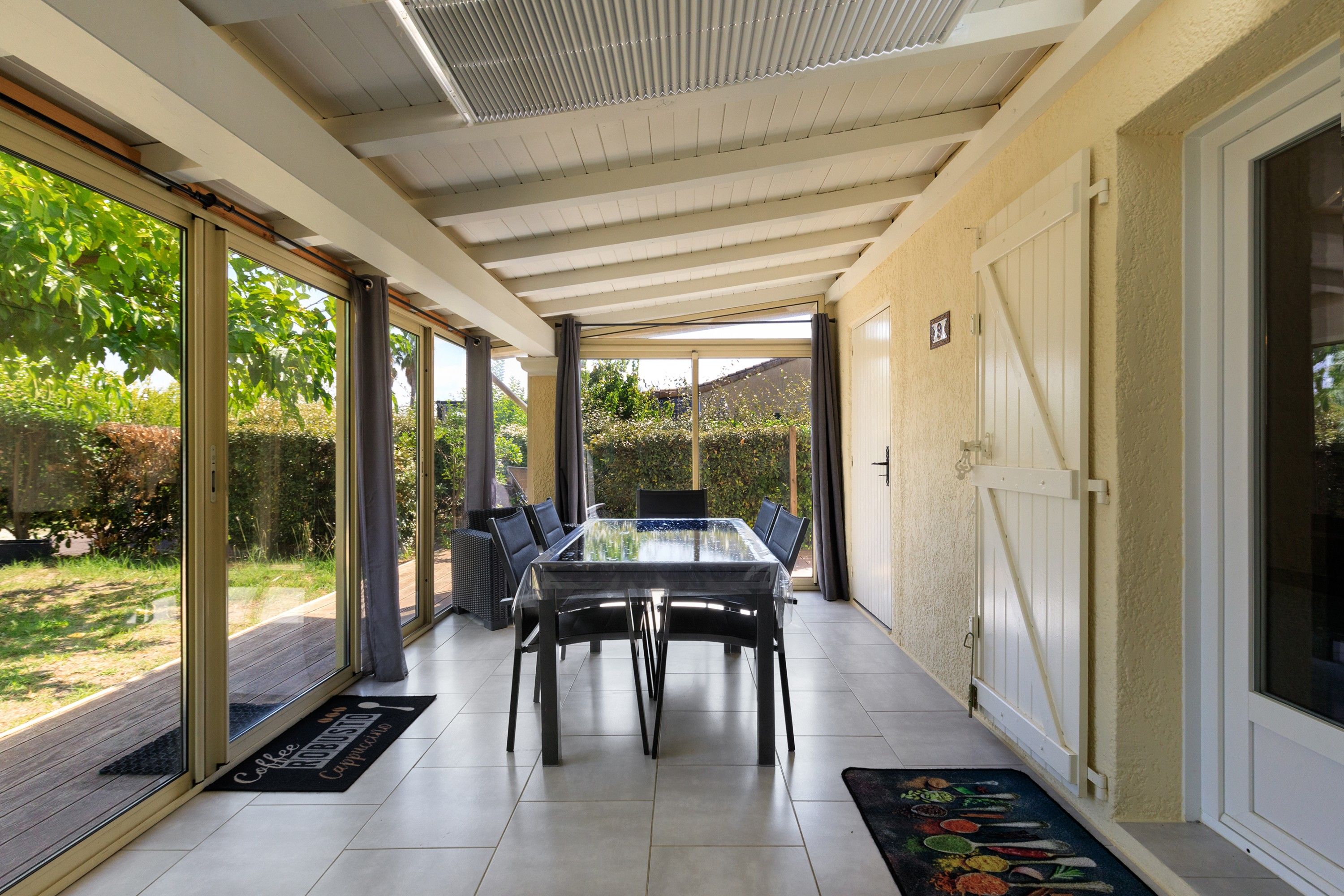 Un patio couvert avec une table et des chaises en verre, une porte et une vue sur la verdure à l’extérieur.