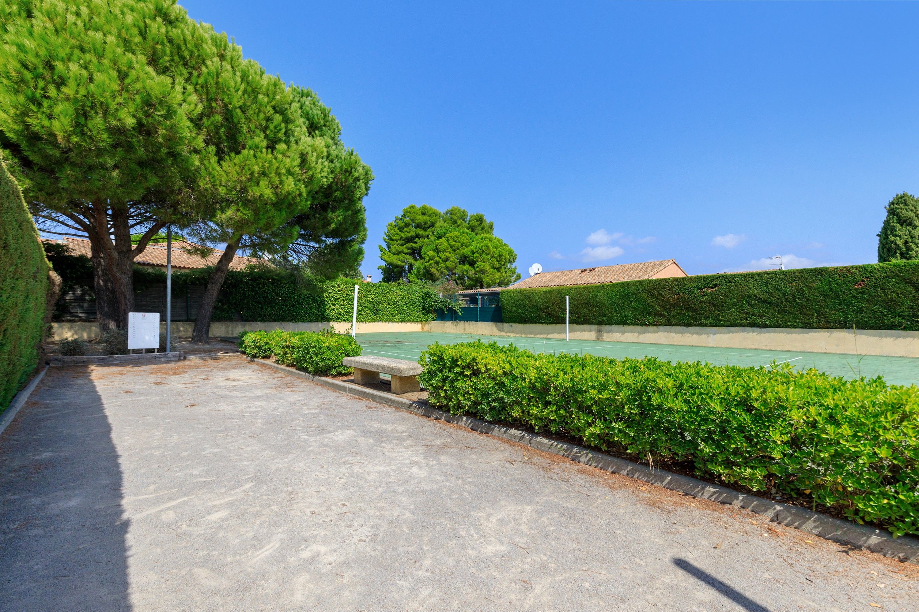 Ein Tennisplatz mit einer Hecke, einer Bank und einem Baum.