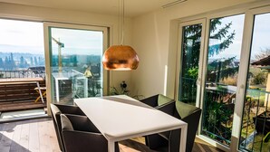 A modern dining area with a white table and dark chairs, large glass doors with a view of a construction site, and a hanging copper pendant light.