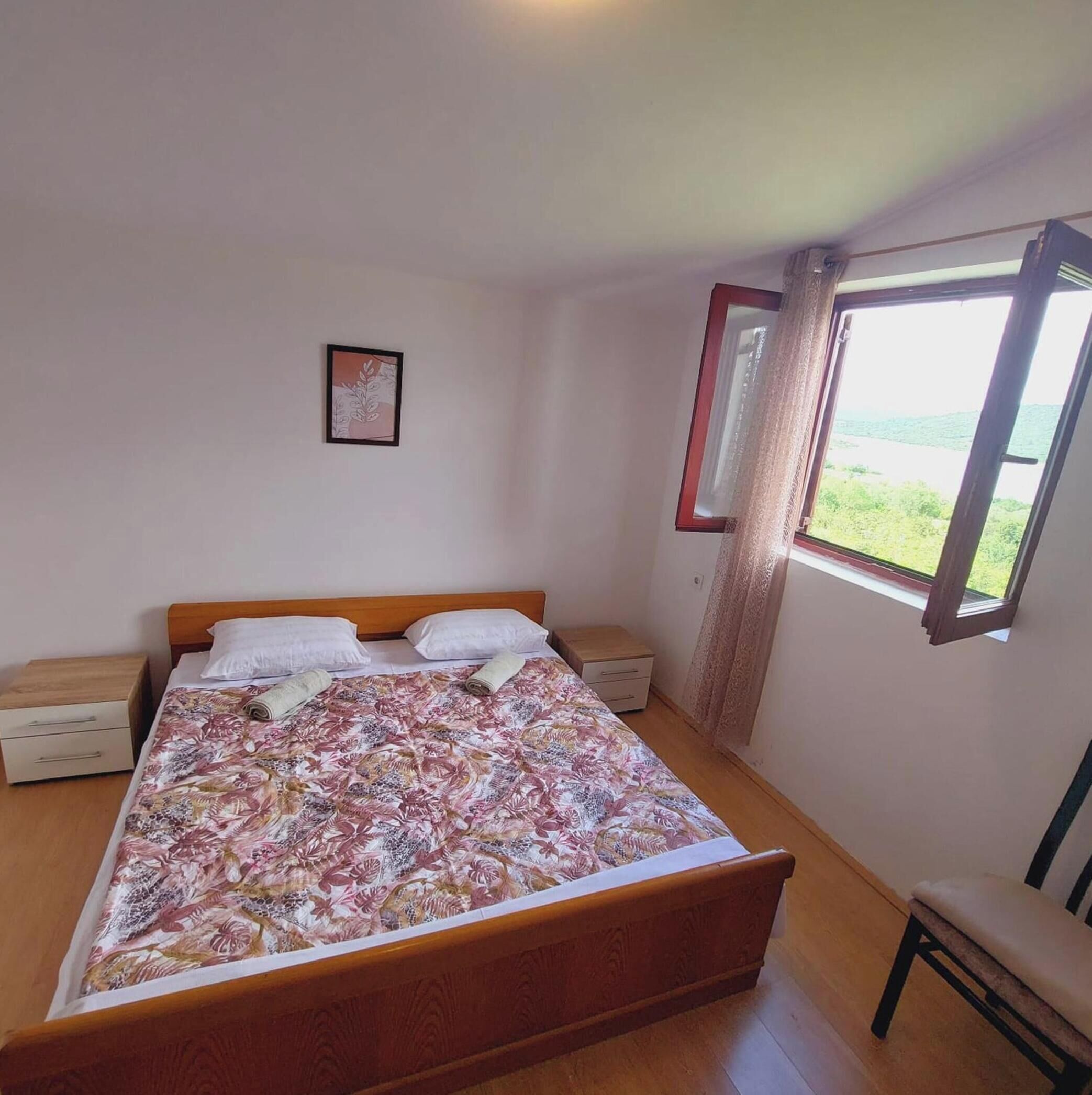 A single bed with a floral bedspread, a nightstand, a small drawer, and a window with a view of greenery.