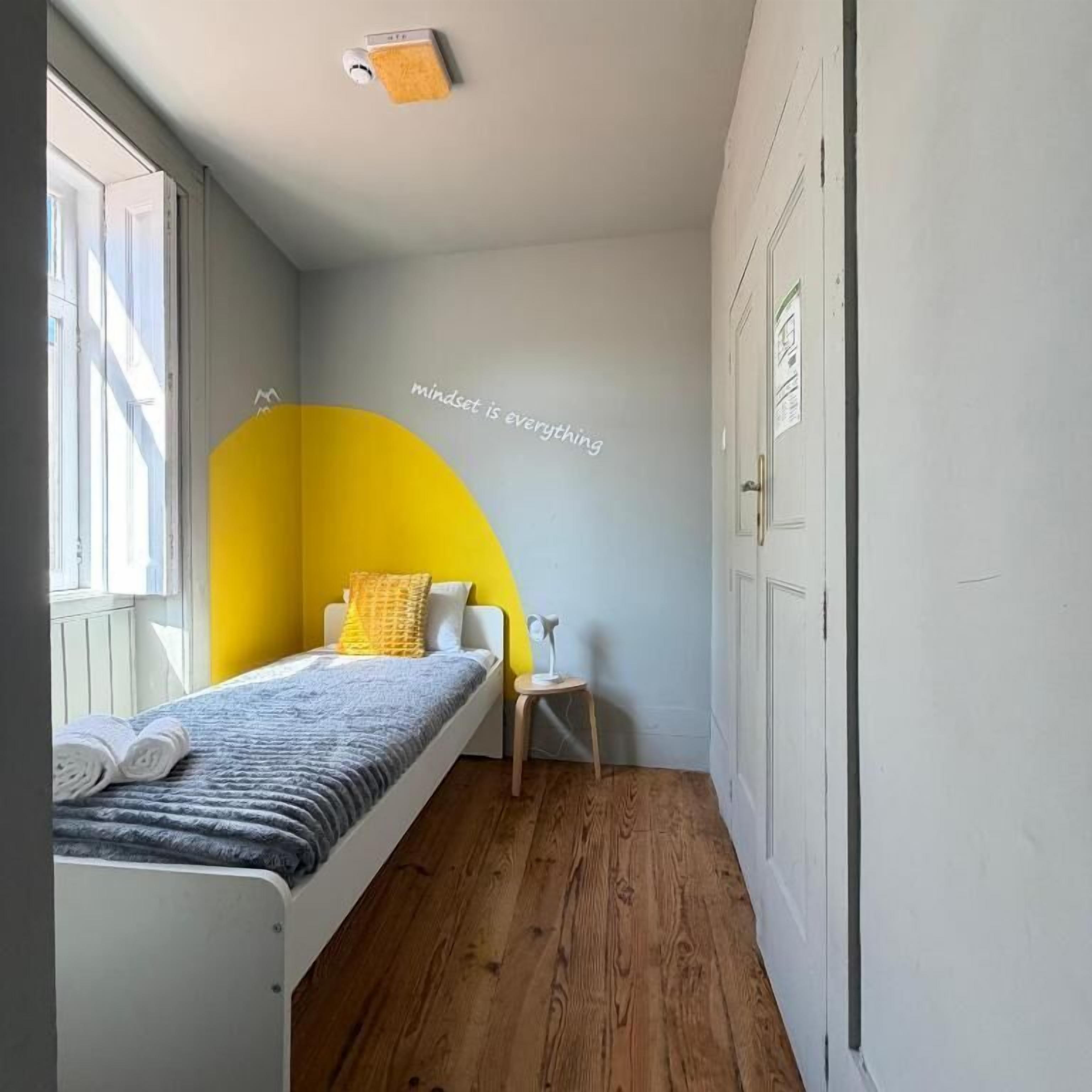 Un dormitorio con cama, una silla, una mesita, un ventanal y una pared con un arco amarillo parcial.