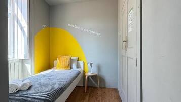Un dormitorio con cama, una silla, una mesita, un ventanal y una pared con un arco amarillo parcial.