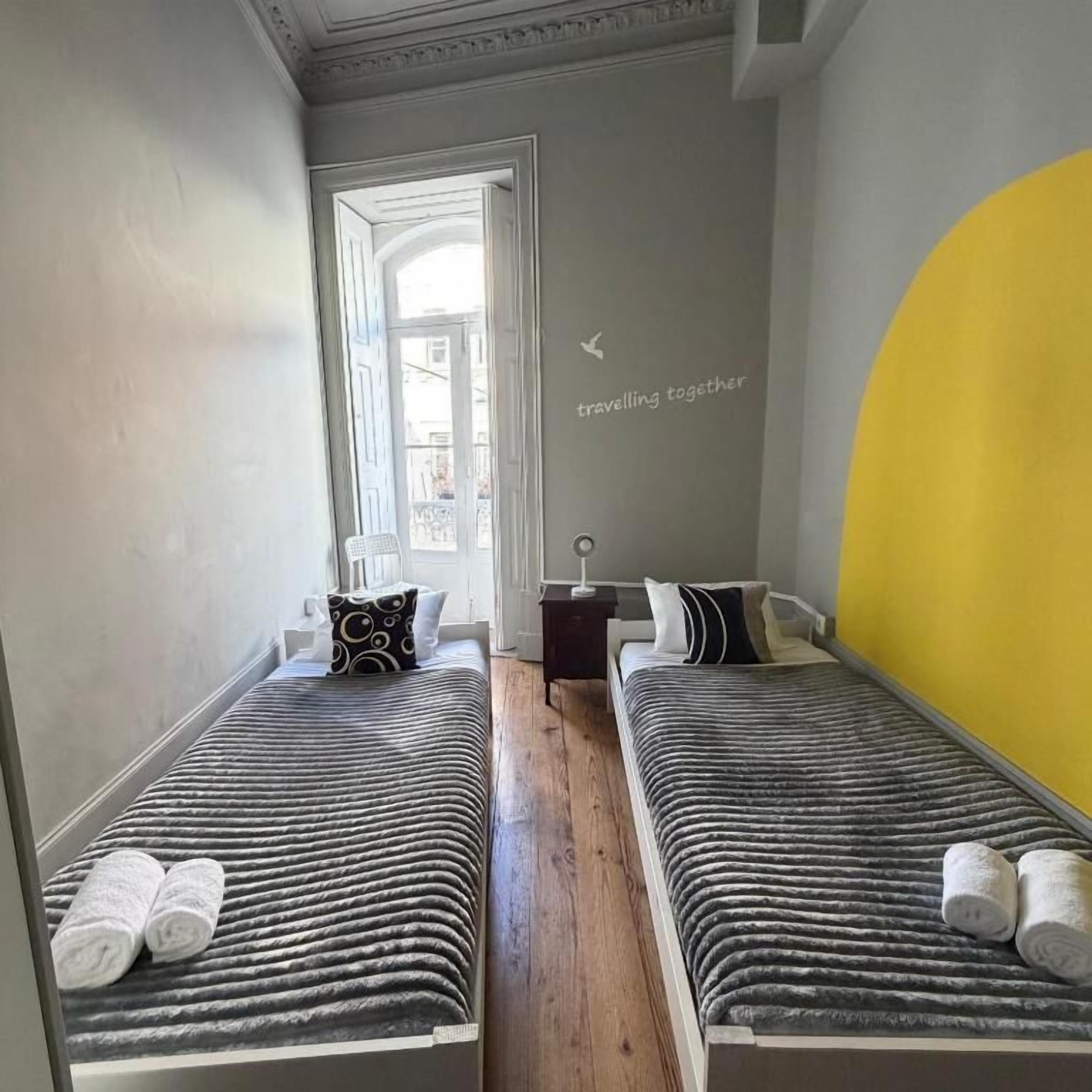 Habitación con dos camas, una mesita pequeña y una silla. Se ve una ventana con vistas al exterior.