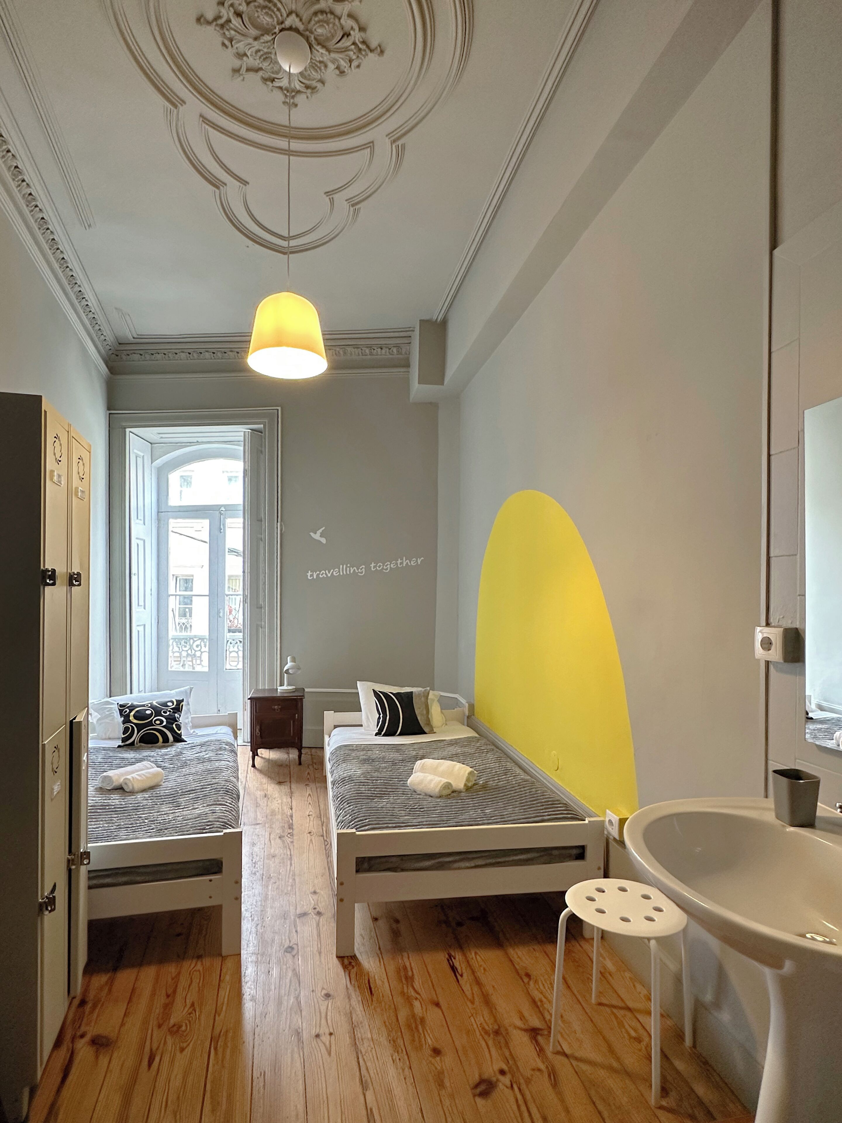 Une chambre avec deux lits, un lavabo, un miroir et une fenêtre donnant sur des immeubles.