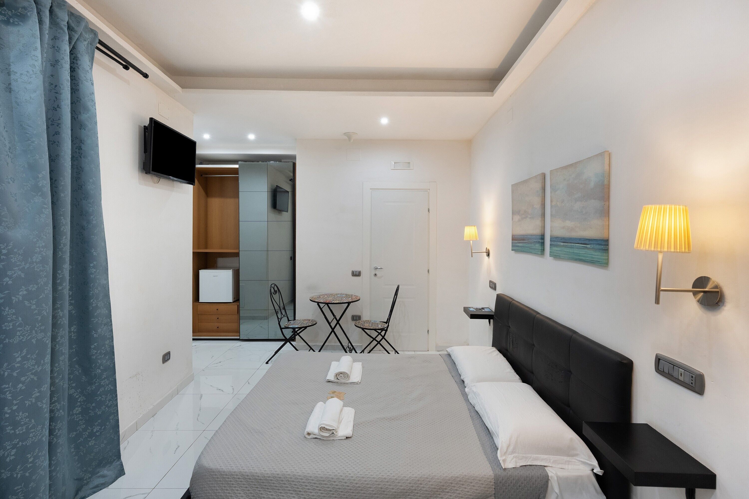 Habitación de hotel con cama, mesita de noche, lámpara, obra de arte y una pequeña mesa con sillas.