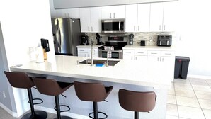 A modern kitchen with a stainless steel refrigerator, a microwave, and a coffee maker. There are three brown bar stools under a white island. - Relaxing House with Gulf Access & Heated Pool (Cape Coral)